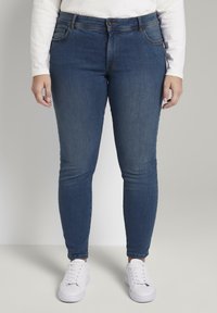 Jeans skinny de mezclilla azul oscuro con cintura media, cinco bolsillos y detalles de costura sutiles, combinados con zapatillas blancas.