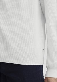 Chemise blanche texturée à manches longues avec un motif en nid d'abeille, présentant un ourlet droit et une petite étiquette de marque sur la couture latérale.