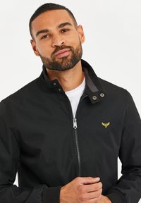 Svart zip-up jacka med grå krage, med en liten gul logotyp på bröstet och ribbade ärmslut. Slät, lättviktsmaterial.