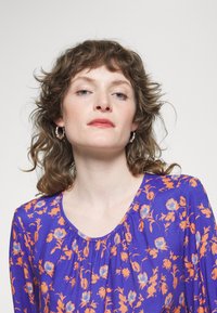 Blusa floral roxa com padrões laranja e azul, decote redondo e detalhes franzidos. Modelo usa brincos de argola prateados.