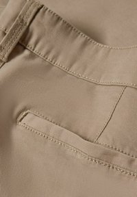 Gros plan sur un tissu beige montrant une poche passepoilée avec des coutures détaillées et une boucle de ceinture sur un vêtement.