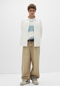 Lässiges Outfit mit einem weißen Hemd, das über einem gestreiften blau-weißen Hemd getragen wird, dazu bequeme beiges Weitgeschnittene Hose und graue Sneakers.