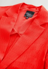 Rød tekstureret blazer med spids krave, enkelt lomme og glat foer. Etiket indeni læser "NEXT." Stoffet har lodrette striber.