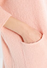Pink knit sweater featuring a textured fabric, long sleeves, and a side pocket with a rounded opening. Soft, cozy appearance.