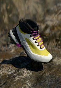 Bota de senderismo amarilla y blanca con detalles en negro, cordones morados y suela resistente. Con puntera reforzada y parte superior texturizada para mayor durabilidad.