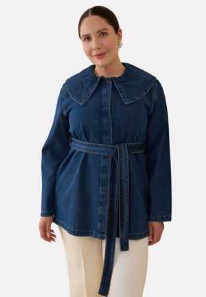 Kvinde med mørkt hår iført en mørk denimjakke med bælte og bred krave samt cremefarvede bukser mod en ensfarvet baggrund.