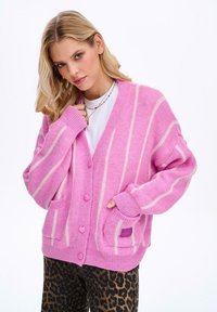 Růžový kardigan s bílými pruhy, knoflíky ve tvaru srdce a předními kapsami. Měkká textura a oversized design. Nosit přes bílý top.