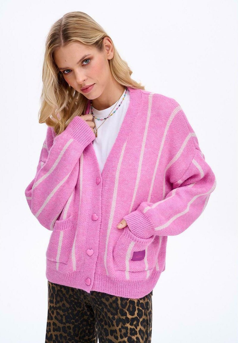 Růžový kardigan s bílými pruhy, knoflíky ve tvaru srdce a předními kapsami. Měkká textura a oversized design. Nosit přes bílý top.