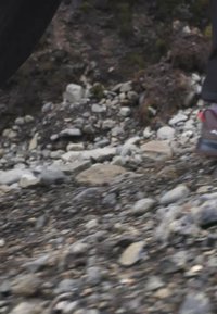 Kaksi jalkaa, joissa mustat housut ja retkeilykengät, kävelee kivikkoisella, epätasaisella maastolla, jossa on pieniä ja suuria kiviä.