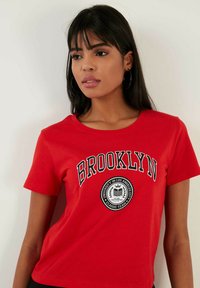 Červené tričko s krátkým rukávem, které na sobě má slovo "BROOKLYN" v černé barvě, kulatý grafický logo a pohodlný výstřih do crew neck.