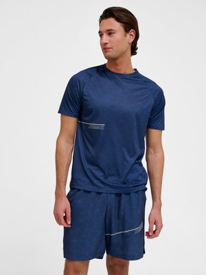 Mann steht und trägt ein marineblaues Sport-T-Shirt mit geometrischem Muster und passende Shorts mit dem "HUMMEL"-Logo, schaut zur Seite.