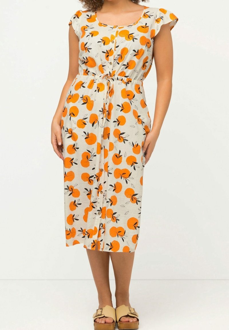Femme portant une robe midi crème avec imprimé fruit orange, manches courtes, boutons sur le devant, taille à cordon et sandales beige à enfiler.