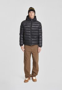Giacca a vento nera con cappuccio, chiusura con zip e design trapuntato testurizzato. Pantaloni marroni e scarpe marroni con lacci bianchi completano l'outfit.