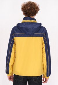 Geltona ir tamsiai mėlyna vėjai atspari striukė su kapišonu, pasižyminti elastingais rankogaliais ir kontrastingomis siūlėmis pečių ir rankovių srityje.