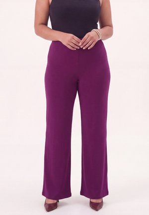 Woman wearing high-waist purple wide-leg pants, black sleeveless top, brown pointed heels, and pearl bracelet, standing with hands clasped.