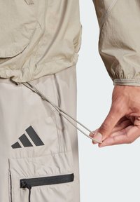 Beige lightweight jacket with a drawstring, elastic cuffs, and a prominent black logo. Features a zippered pocket and soft texture.
