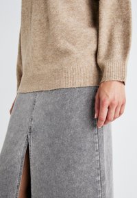 Bež pleteni pulover z rebrastimi manšetami v kombinaciji s sivo jeans krilo z bočnim razporkom, ki prikazuje mehko teksturo in sproščen dizajn.
