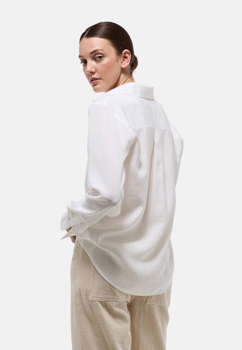 Chemise blanche à manches longues avec col et poignets boutonnés. Tissu doux et lisse avec une coupe décontractée, associée à un pantalon beige texturé.