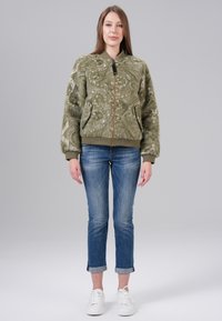 Veste bomber vert olive avec une texture douce et un motif discret, associée à un jean bleu retroussé et des baskets blanches.
