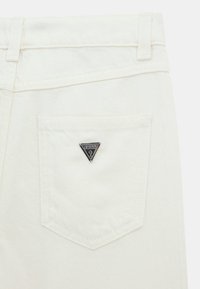 Jeans in denim bianco con un logo triangolare nero "Guess?" sulla tasca posteriore, caratterizzati da una forma standard della tasca e una texture sottile.