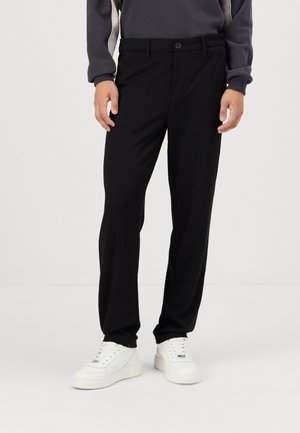 Mann trägt schwarze, gerade geschnittene Hose, weiße Sneakers und ein dunkelgraues langärmeliges Oberteil und steht vor einem einfarbigen weißen Hintergrund.