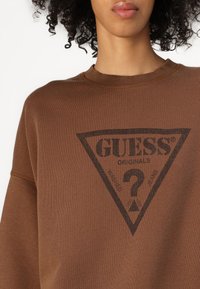 Barna pulóver bordázott nyakkivágással, középen fekete háromszög logóval és "GUESS ORIGINALS" felirattal. Puha szövet textura.