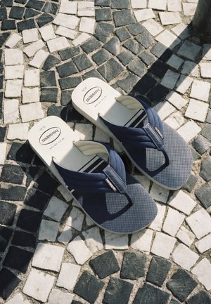 Paire de tongs Havaianas bleu marine et blanc sur un pavé carrelé irrégulier noir et blanc sous le soleil et l'ombre.