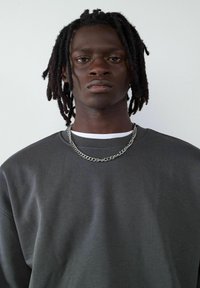 Szary sweter z okrągłym dekoltem, z teksturowaną powierzchnią. Srebrny naszyjnik na łańcuszku dodaje szczegółu do dekoltu. Widoczna prosta biała podkoszulka.