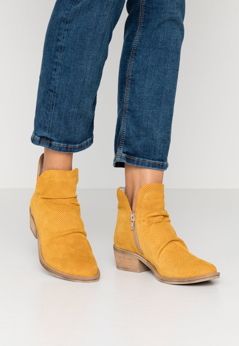 Bottines en daim jaune avec fermetures éclair latérales portées avec un jean bleu retroussé sur un fond clair.
