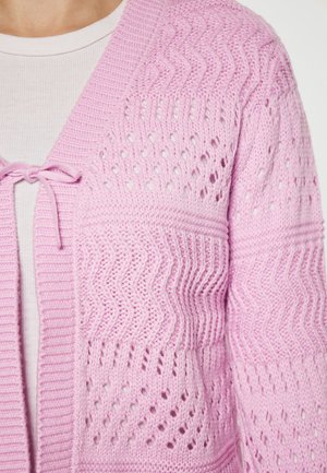 Cardigan tricotat de culoare roz deschis, cu o textură ondulată, model cu ochiuri deschise și închidere cu funda la gât.