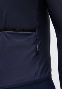 Κοντινό πλάνο στην πλαϊνή τσέπη ενός ναυτικού μπλε μπουφάν με μαύρο φερμουάρ που φέρει την ετικέτα "GripGrab" και μια υφασμάτινη μαύρη λωρίδα πάνω από την τσέπη.