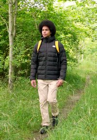 Svart puffermantel, beige byxor och svarta stövlar med en gul ryggsäck. Omgiven av grönska på en lerig stig.