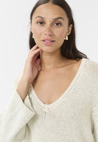 Pull en tricot de couleur crème clair, avec un décolleté en V profond, des manches longues, et un motif texturé, avec une coupe ample et des détails d'ourlet brut.