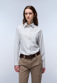 Striped button-up shirt in light blue and white, made of fabric with a smooth texture. Paired with khaki pants and a brown leather belt.