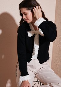Black knitted cardigan with white lace trim at collar and cuffs, featuring a bow at the neck. Model pairs it with cream trousers.