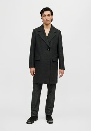 Un homme se tient devant un fond uni et clair, vêtu d’un long manteau noir, d’un pantalon foncé et de chaussures noires.
