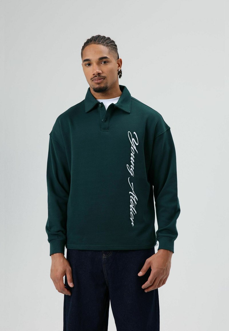 Donkergroen poloshirt van katoen, met een zijlogo in witte letters. Lange mouwen met een losse pasvorm, kraag met revers.