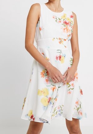 Frau trägt ein ärmelloses weißes Kleid mit mehrfarbigem Blumenmuster und tailliert, steht mit verschränkten Händen.