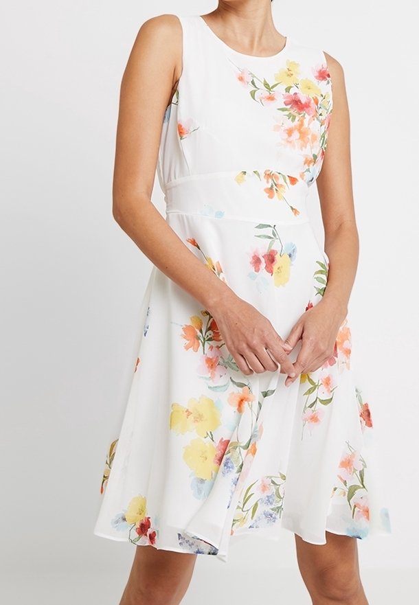 Femme portant une robe blanche sans manches avec un imprimé floral multicolore et une taille ajustée, debout, les mains jointes.