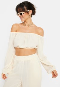 Cremfarbener Crop-Top mit schulterfreien Ärmeln, fließenden Ärmeln und strukturiertem Stoff. Kombiniert mit passenden Hosen, die einen elastischen Bund haben.