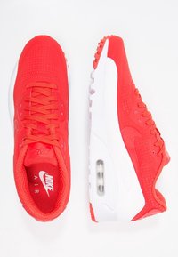 Des sneakers Nike Air rouge vif avec des semelles blanches, un amorti d'air visible et des lacets détaillés, présentées en vues de dessus et de côté sur un fond blanc.