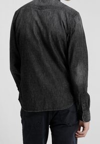 Mörkgrå denimskjorta med lång ärm, rundad nederkant och knapprad på ärmsluten. Tyget har en subtil textur och slitageuttryck.