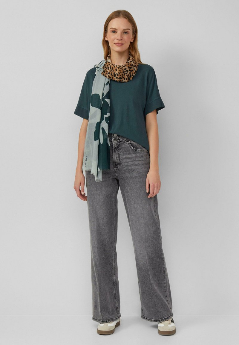 Teal T-shirt met korte mouwen, gecombineerd met ruime grijze jeans. Een gestreepte beige en zwarte luipaardprint sjaal hangt om de nek.