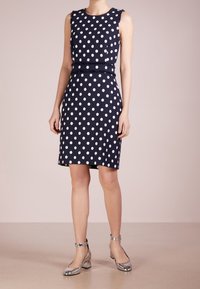 Námořnická modrá bezramínková šaty s bílými puntíky, vyrobené z texturované látky, s páskem v pase. Modelka má na sobě stříbrné sandály na kotníku.