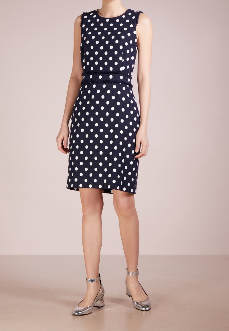 Námořnická modrá bezramínková šaty s bílými puntíky, vyrobené z texturované látky, s páskem v pase. Modelka má na sobě stříbrné sandály na kotníku.