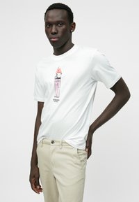 T-shirt blanc à manches courtes avec un graphisme d'une canette rose entourée de flammes, assorti à un pantalon beige. Design simple, en coton.