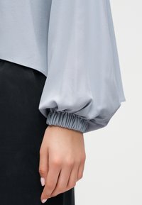 Main à carnation claire portant un chemisier à manches longues gris clair avec poignets élastiques froncés, sur un fond clair uni.