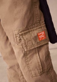 Beiga cargobyxor tillverkade av hållbart material, med en stor ficka med orange Superdry-logotyp och synliga sömmar.