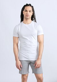 Νέος άνδρας με μακριά dreadlocks, φοράει εφαρμοστό λευκό μπλουζάκι και γκρι σορτς, στέκεται μπροστά σε απλό ανοιχτό φόντο.