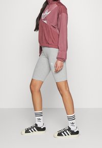Maroon zip-up jacka, grå cykelshorts, svartvita sneakers, randiga strumpor, med Adidas-logotyper och en slät, sportig design.
